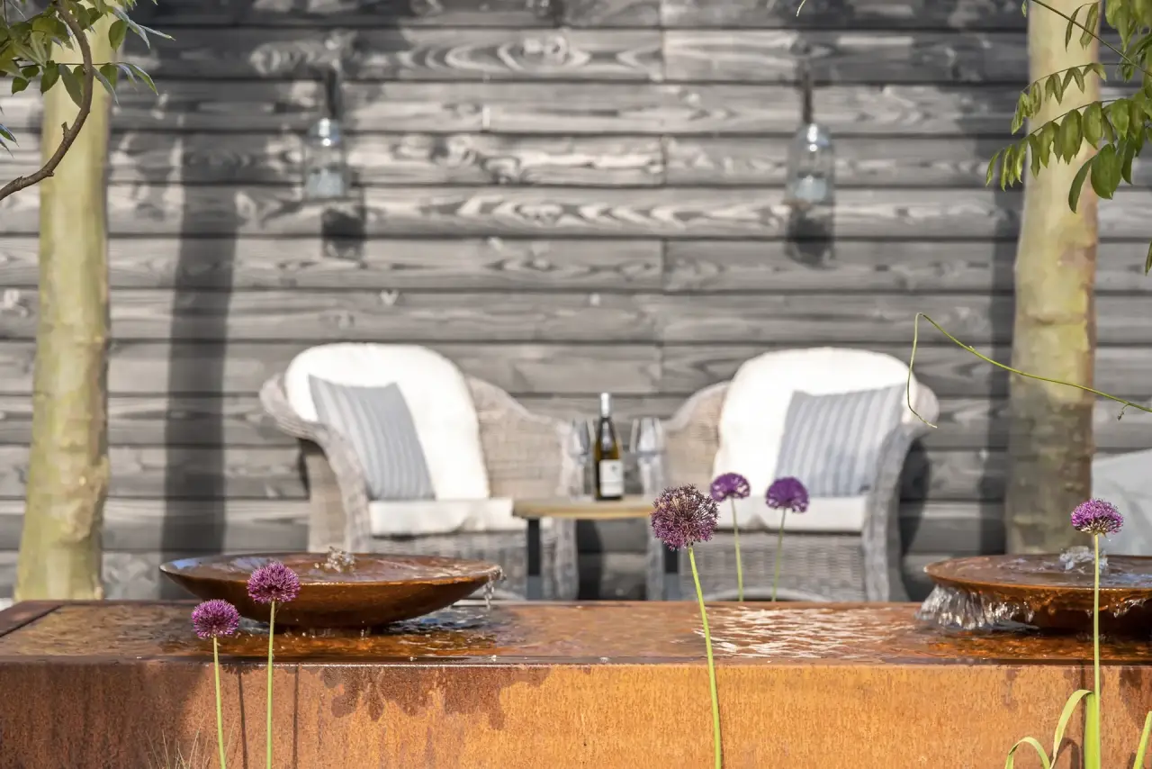 Cortenstalen vijverbak met twee stoelen ernaast tegen een houten wand.
